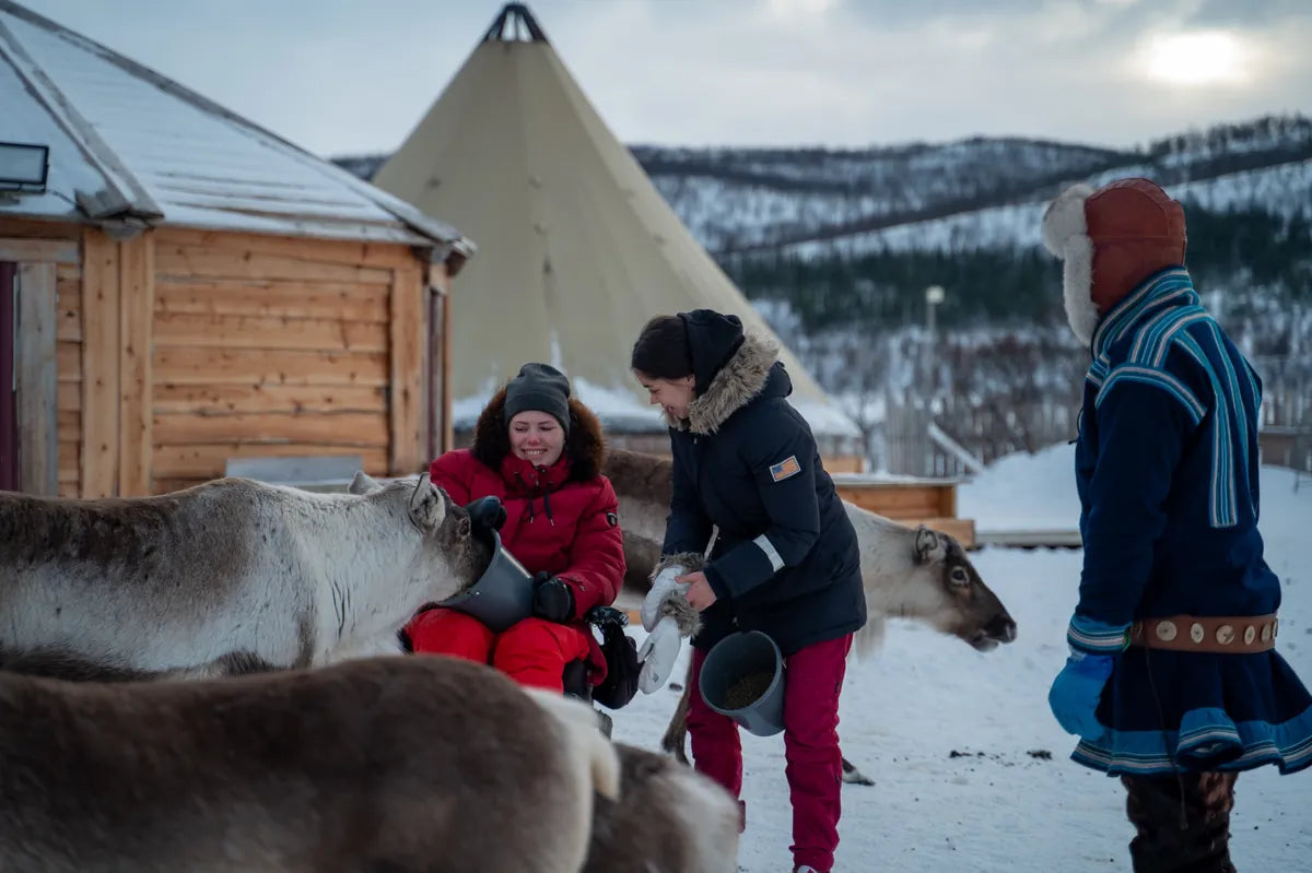 Two people interacting with reindeer in a snowy landscape with traditional huts and mountains in the background.